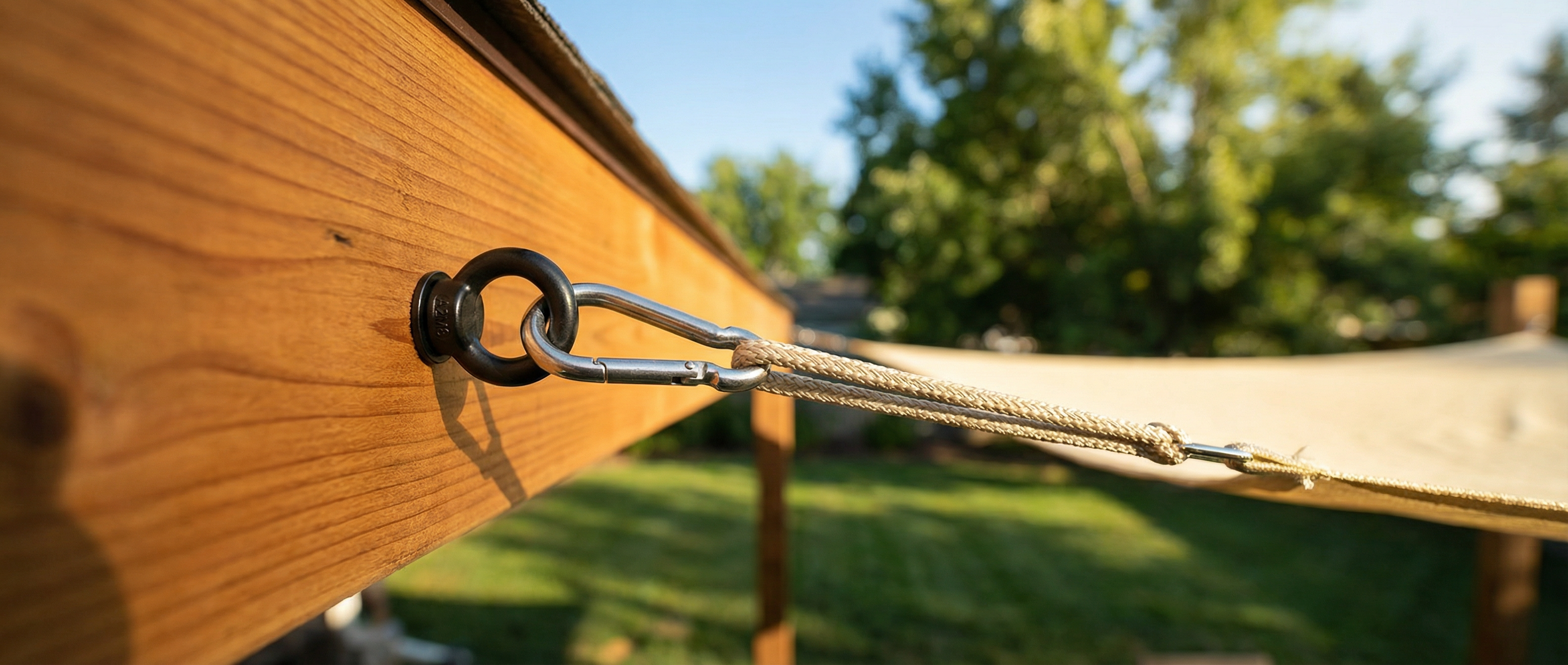 Close-up of a heavy-duty black stainless steel eye bolt installed on a wooden pergola beam. The rust-proof hardware connects to a silver snap hook and rope to securely anchor a beige sun shade sail over a sunny backyard patio.