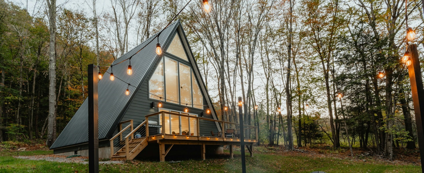 A cozy wooden A-frame cabin in a forest clearing at dusk, featuring warm Edison-style outdoor string lights suspended tautly across the yard with a heavy-duty wire rope hanging kit.